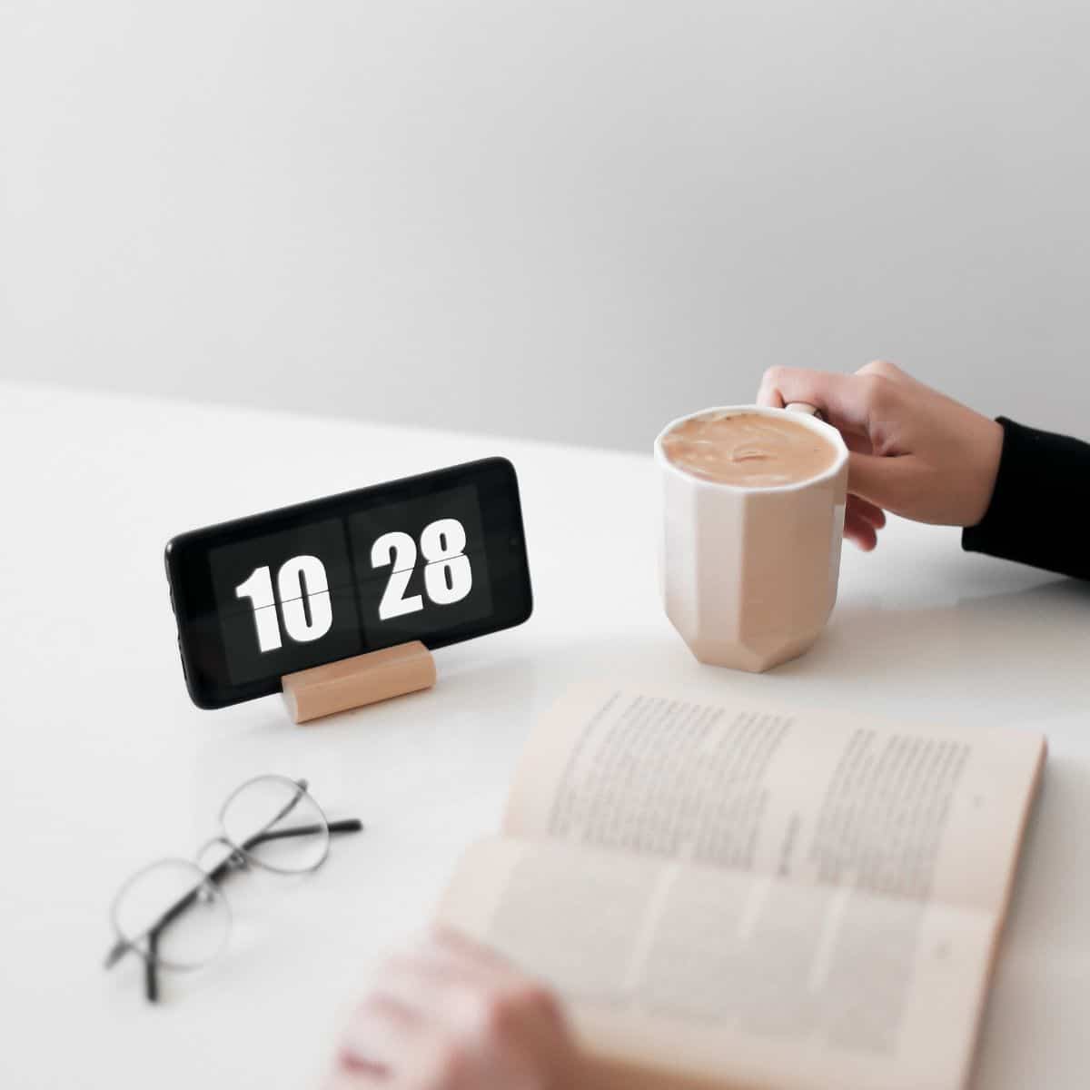 person reading a book near a clock with a cup of coffee.