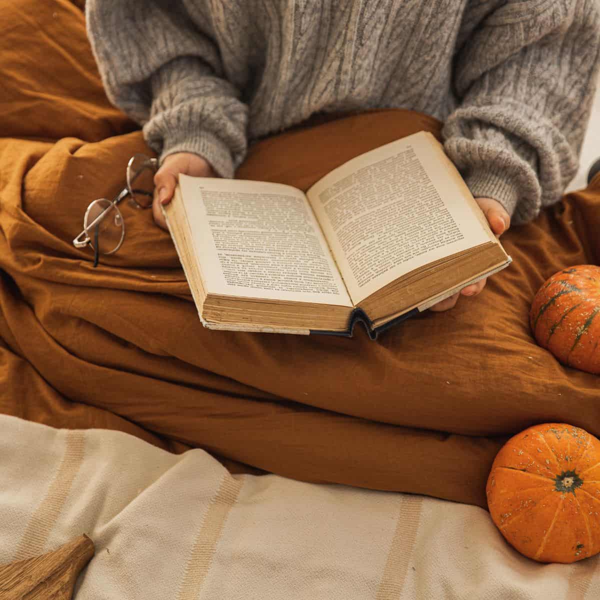 woman sitting under a blanket reading a book near pumpkins.