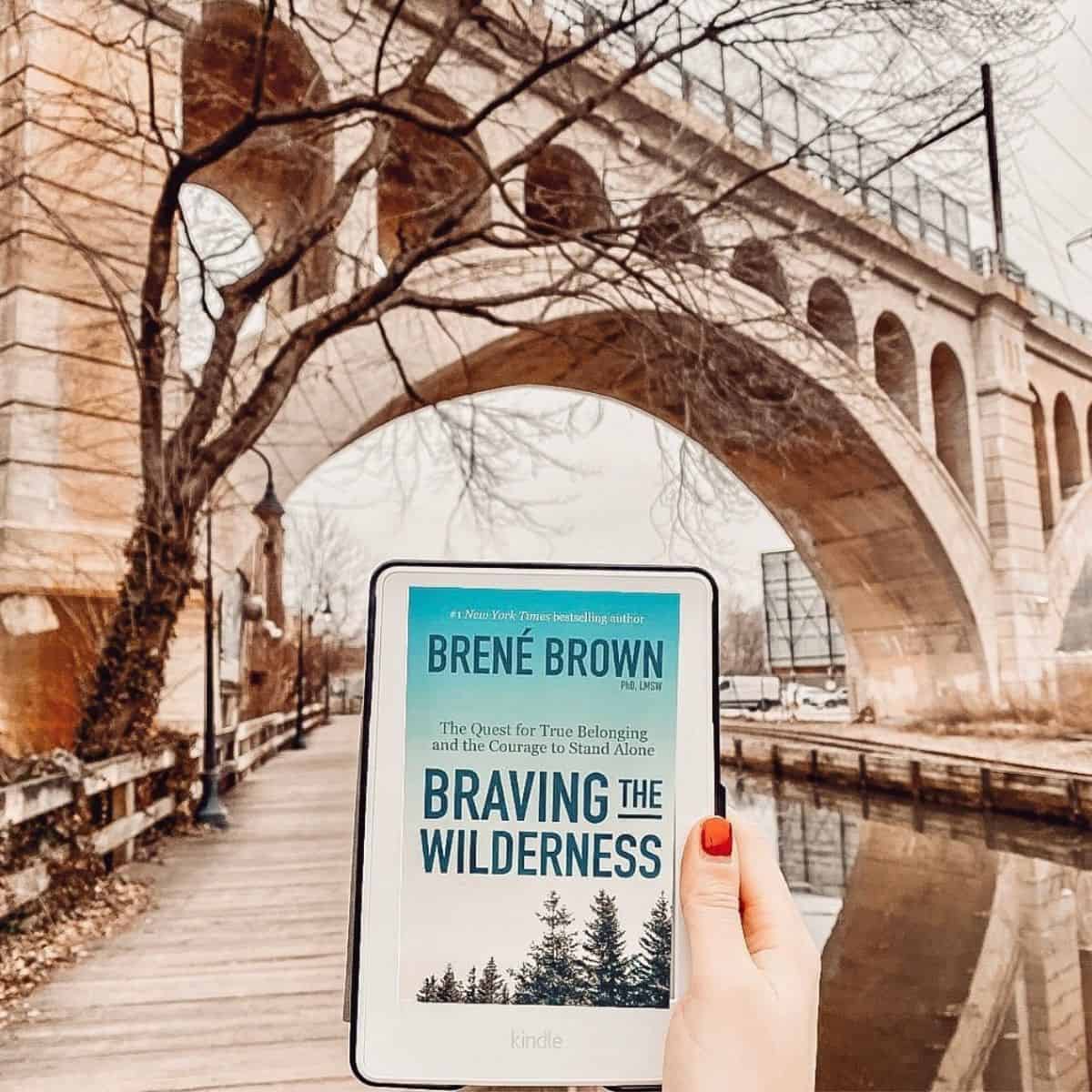 person holding braving the wilderness by brene brown in front of a bridge.