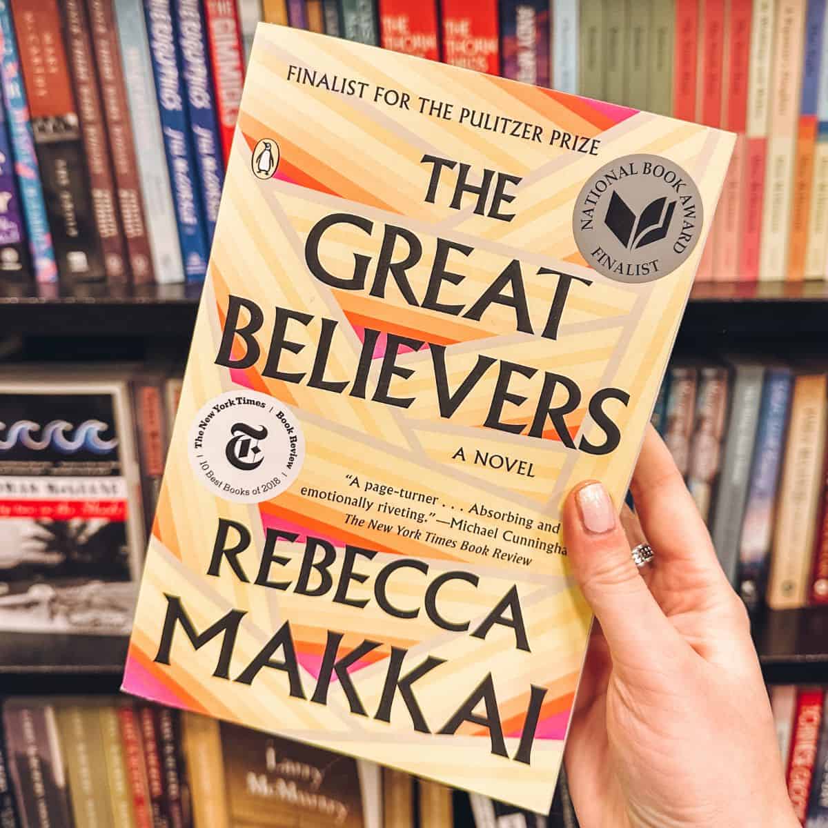 The great Believers by Rebecca Makkai held in front of a bookshelf.